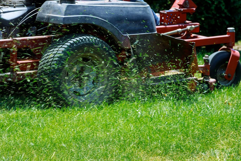 A riding mower in action cutting grass with blades of grass flying around.